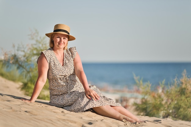 Mulher europeia feliz com vestido longo e chapéu de palha senta-se na praia e desfruta de férias à beira-mar