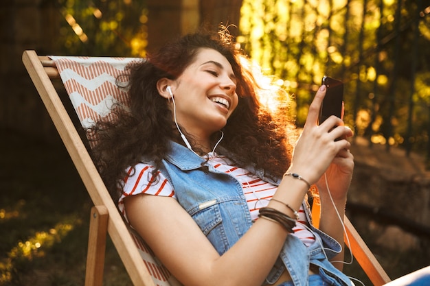 mulher europeia feliz rindo e ouvindo música com fones de ouvido, enquanto está sentado na espreguiçadeira durante o descanso no parque em um dia ensolarado