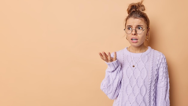 Mulher europeia indignada descontente com cabelo loiro penteado parece com expressão infeliz mantém a palma levantada vestida com poses de jumper de malha contra fundo marrom espaço em branco para sua promoção