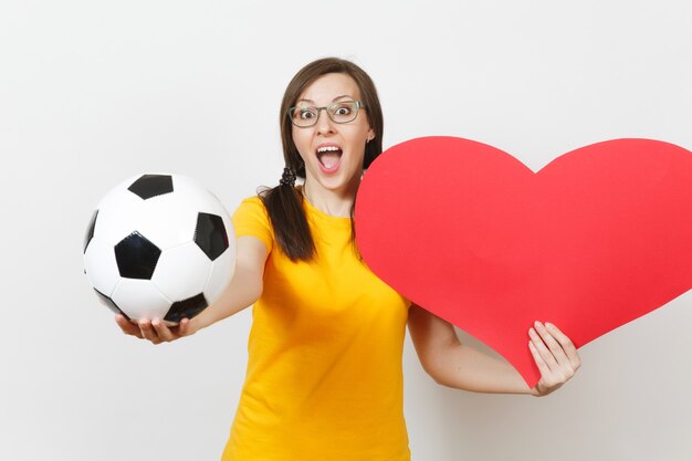 Mulher europeia sorridente, divertido rabo de cavalo, fã de futebol ou jogador de óculos, uniforme amarelo segura bola de futebol clássica, coração vermelho isolado no fundo branco. Esporte, futebol, conceito de estilo de vida saudável.