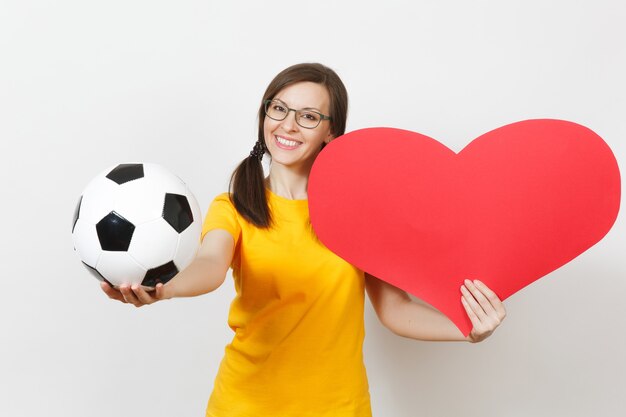 Mulher europeia sorridente, divertido rabo de cavalo, fã de futebol ou jogador de óculos, uniforme amarelo segura bola de futebol clássica, coração vermelho isolado no fundo branco. Esporte, futebol, conceito de estilo de vida saudável.