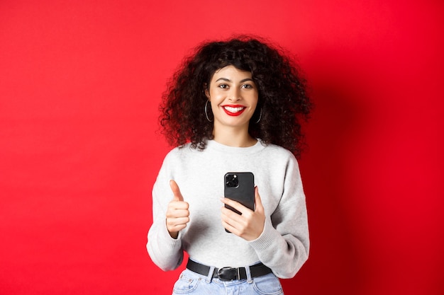 Mulher europeia sorridente, parecendo feliz e satisfeita, usando o celular e fazendo o polegar para cima, recomendando o aplicativo, em pé na parede vermelha.