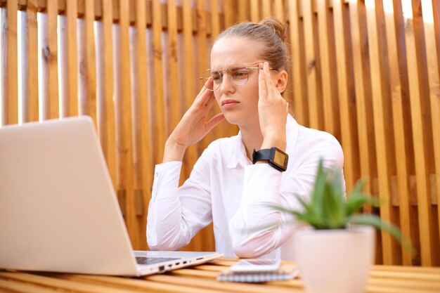Mulher exausta cansada vestindo camisa branca sentada no café ao ar livre trabalhando no laptop com dor de cabeça sendo sobrecarregada franzindo a testa massageando os templos
