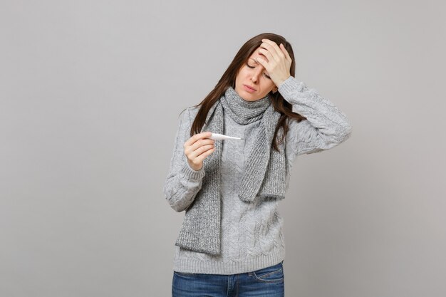 Foto mulher exausta de suéter, lenço colocado a mão na testa, espera olhando no termômetro isolado no fundo cinza. estilo de vida saudável, tratamento de doença doente, conceito de estação fria. simule o espaço da cópia.
