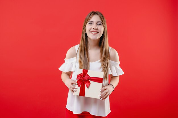 Mulher excitada com caixa de presente, vestindo blusa branca isolada na parede vermelha