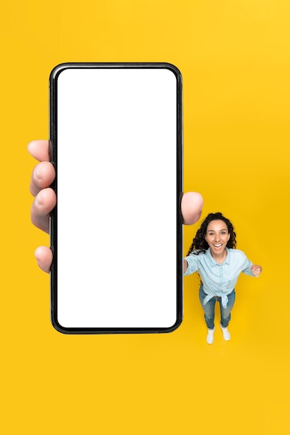 Mulher excitada mostrando tela enorme de telefone sobre fundo amarelo vertical