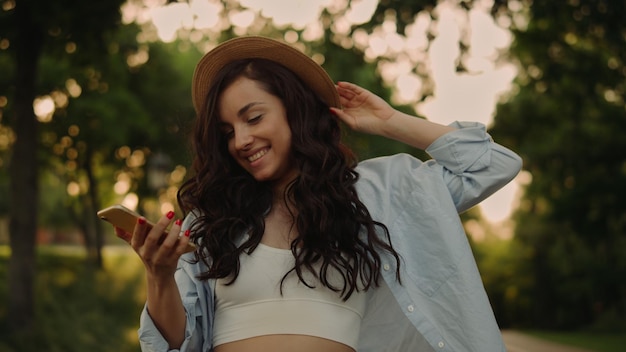 Mulher excitada usando telefone celular no parque Mulher positiva andando com smartphone