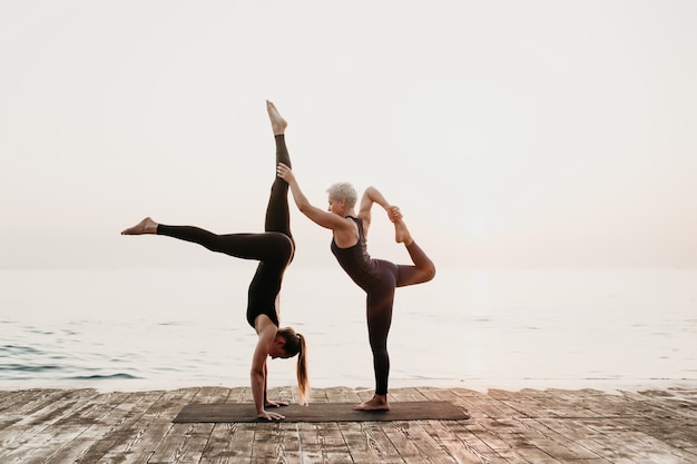 Mulher exercitando acro yoga na praia perto do mar pela manhã com o tutor feminino maduro