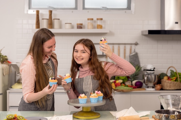 Mulher expectante e filha decorando cupcakes em uma mesa da cozinha, se divertindo enquanto comem pastéis recém-assados