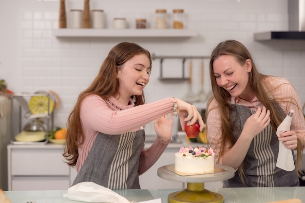 Mulher expectante e filha decorando cupcakes em uma mesa da cozinha, se divertindo enquanto comem pastéis recém-assados