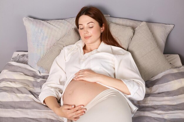 Mulher expectante satisfeita pacificamente tirando uma soneca na cama de casa vestindo camisa branca acariciando suavemente sua barriga e mantendo os olhos fechados aproveitando seu descanso