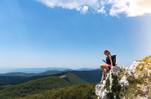 Mulher exploradora sentada olhando seu smartphone com uma vista panorâmica da montanha ao fundo