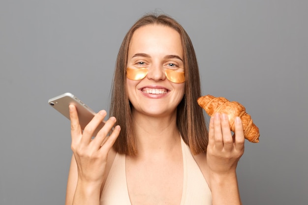 Mulher extremamente feliz com cabelo castanho em pé com croissant e telefone inteligente olhando para a câmera com um sorriso de dentes usando o telefone móvel em pé com manchas sob os olhos