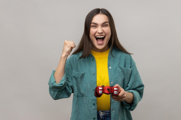 Mulher extremamente feliz segurando a almofada do joystick vermelho de mão mostrando sim gesto sinceramente regozijando-se com a vitória no jogo virtual online vestindo jaqueta estilo casual Tiro de estúdio interno isolado em fundo cinza