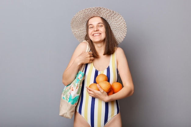 Mulher extremamente feliz vestindo trajes de banho listrados e chapéu de palha em pé com laranjas frescas nas mãos, satisfeita com dias quentes de descanso tropical perto do mar isolado sobre fundo cinza