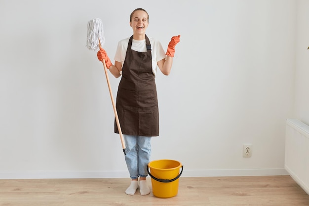 Mulher extremamente satisfeita usando luvas de borracha laranja, avental marrom e jeans lavando o chão em casa, segurando o esfregão e os punhos cerrados, feliz em terminar de limpar a casa.