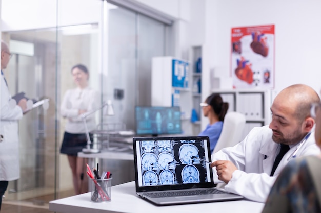 Mulher falando com o médico durante a consulta sobre raio-x cerebral usando laptop no hospital. Médico visitante sênior do paciente, relatando queixas de saúde, e o médico explicando o tratamento.