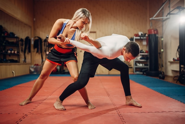 Mulher faz chute de cotovelo, treino de autodefesa com personal trainer masculino, interior do ginásio. Mulher em treinamento, prática de defesa pessoal