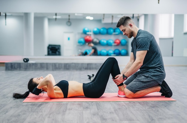 Mulher fazendo abdominais, pressione o exercício no tapete com seu treinador esportivo masculino na academia Exercícios de casal no fundo desfocado da academia