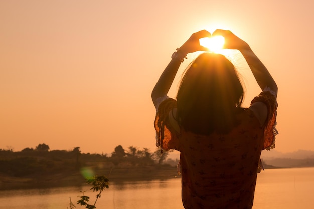 Foto mulher fazendo forma de coração na beira do lago contra o céu claro durante o pôr do sol