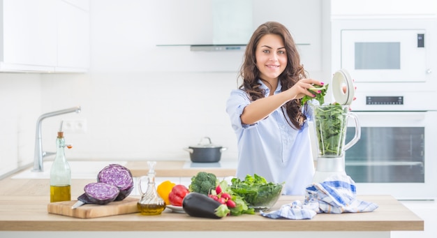 Mulher fazendo sopa de vegetais ou smoothies com o liquidificador na cozinha dela. Jovem mulher feliz preparando uma bebida saudável com rúcula de brócolis espinafre e outros vegetais.