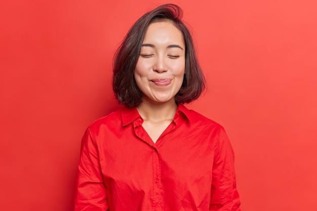 mulher fecha os olhos lambe os lábios da tentação de provar algo delicioso mostra a língua imagina comer comida deliciosa usa camisa isolada em vermelho vivo