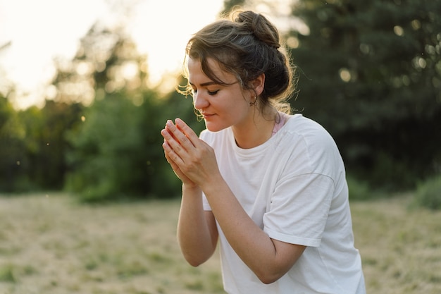 Mulher fechou os olhos orando em um campo durante o belo pôr do sol com as mãos postas em conceito de oração para ...