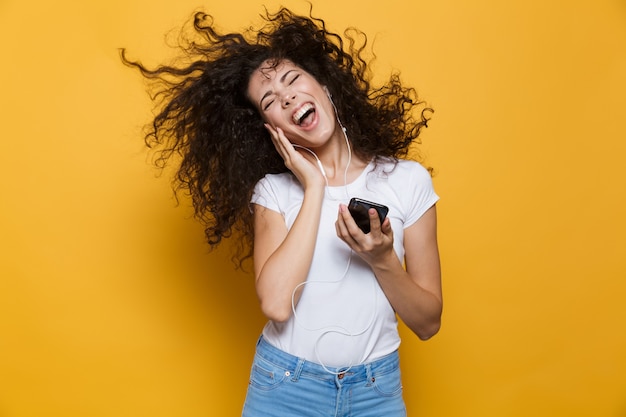 mulher feliz 20 anos com cabelo ondulado e ondulado cantando enquanto segura o smartphone e ouve música com fones de ouvido