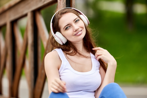 Mulher feliz 29 anos em fones de ouvido sem fio está sentada em um parque público no dia de verão.