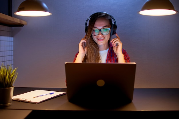 Mulher feliz, alegre, satisfeita e sorridente, usando fones de ouvido sem fio, desfrutando de assistir um filme online usando um laptop à noite