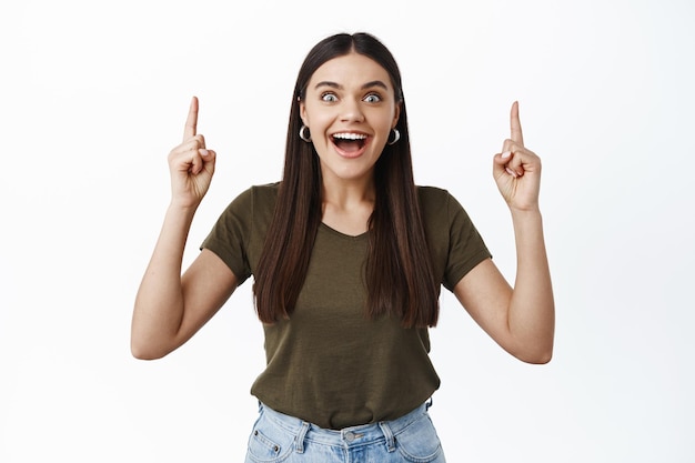 Foto mulher feliz animada com longos cabelos escuros, boca aberta fascinada, apontando os dedos para cima, em pé sobre uma parede branca