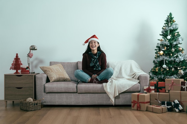 Mulher feliz aproveitando o tempo de lazer no Natal relaxando no sofá pela árvore de Natal com um sorriso radiante. jovem garota asiática alegre com chapéu de Papai Noel vermelho e suéter rindo câmera de rosto sentado no sofá copyspace