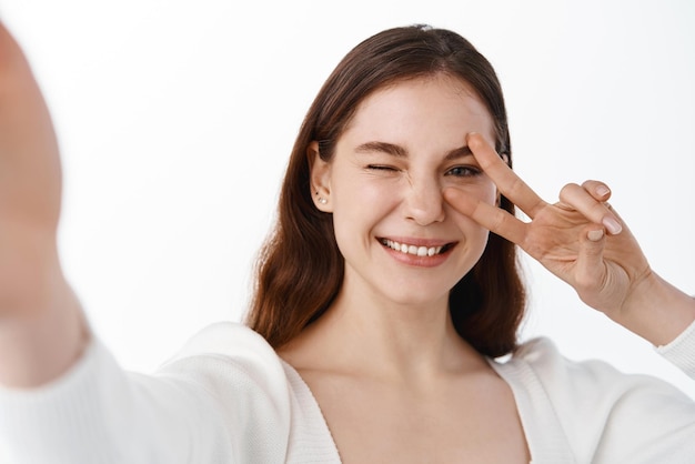 Mulher feliz atraente tomando selfie com vsign estende a mão para segurar a câmera fazendo um sorriso de rosto positivo e gesto de paz em pé sobre fundo branco