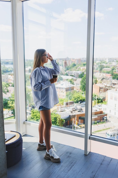 Mulher feliz bebe café e olha pela janela em um café Uma jovem sorridente de óculos está de pé junto à janela Lazer de lazer e tempo livre O estilo de vida freelancer moderno