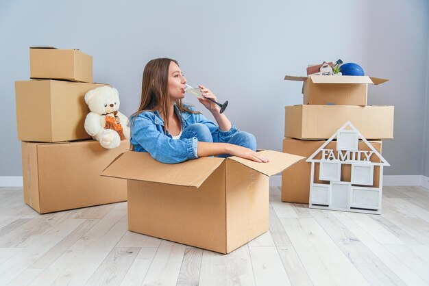 Mulher feliz bebe champanhe de um copo enquanto está sentada dentro de uma caixa de papelão em um novo apartamento