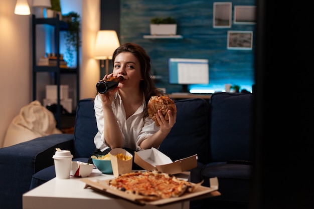 Foto mulher feliz bebendo cerveja, comendo hambúrguer saboroso e delicioso assistindo filme da série de documentários