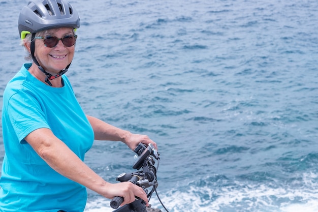 Mulher feliz ciclista maduro que aprecia a atividade no mar com sua bicicleta elétrica. De pé no penhasco