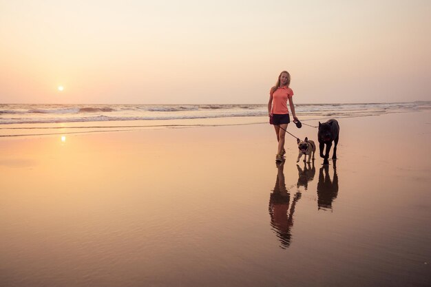 Mulher feliz com bulldog francês e cone corso caminhando e brincando na praia de goa índia ao pôr do sol