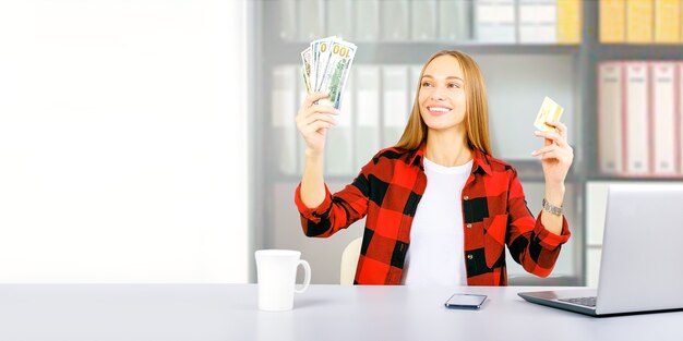 Mulher feliz com cartão de crédito e notas de dólar com uma xícara de café e seu laptop sobre uma mesa branca