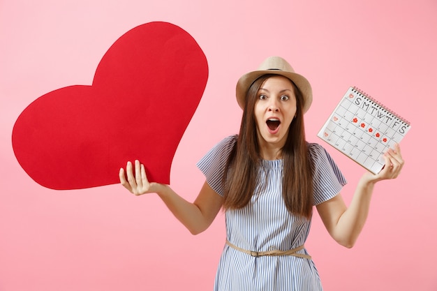 Mulher feliz com chapéu de verão vestido azul segurando coração vermelho grande em branco vazio, calendário de períodos femininos, verificando os dias de menstruação isolados no fundo. Conceito ginecológico de saúde médica. Copie o espaço