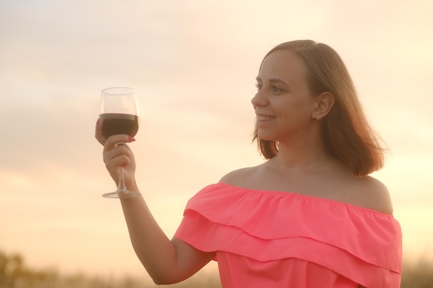 Mulher feliz com copo de vinho na zona rural Vista lateral da jovem fêmea de vestido vermelho em pé com copo de vinho tinto e desviar o olhar
