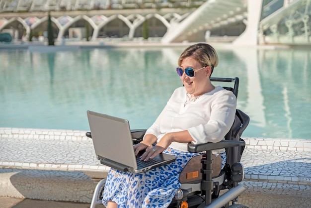 Mulher feliz com deficiência sentada em cadeira de rodas usa laptop fora para trabalho remoto