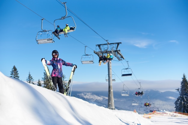 Mulher feliz com esquis em pé perto de elevador na encosta da montanha coberta de neve. Dia de sol durante as férias de inverno. Visão geral.