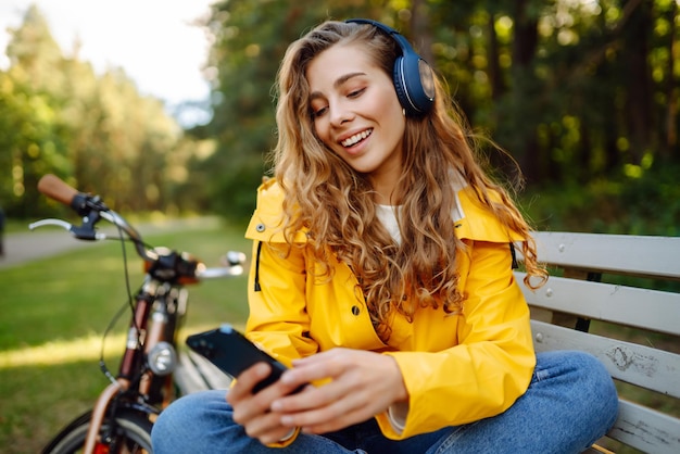 Mulher feliz com fones de ouvido, smartphone, andar de bicicleta num parque ensolarado, estilo de vida ativo, descanso