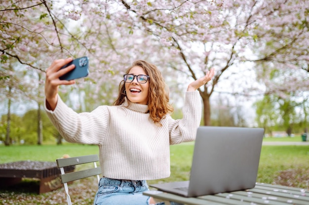 Mulher feliz com laptop e telefone no parque de flores de primavera enquanto trabalha on-line conceito de freelance