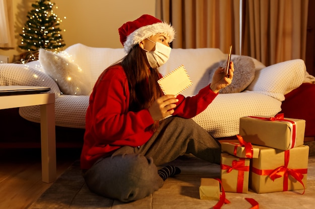 Mulher feliz com máscara facial sozinha em casa, comemorando o Natal com videochamada online para família