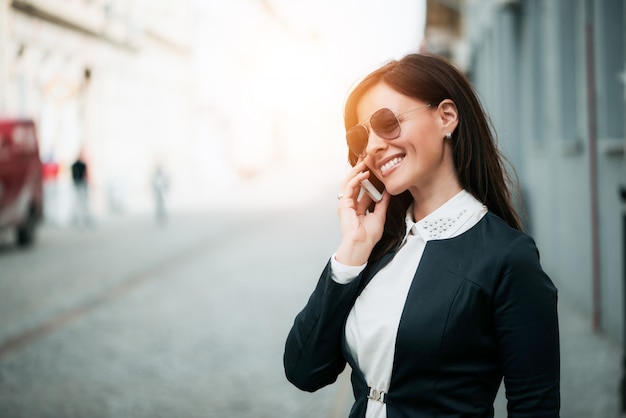 Mulher feliz com óculos de sol sorrindo brilhantemente enquanto fala no telefone