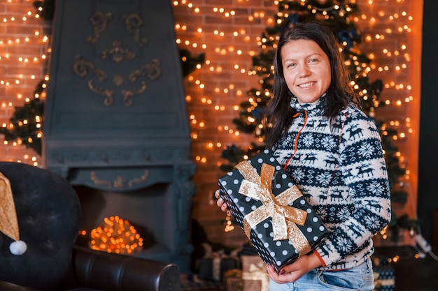 Mulher feliz com roupas de natal com caixa de presente em pé na sala decorada de ano novo.