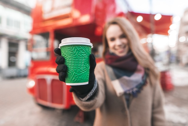 Mulher feliz com um casaco em pé na rua segurando nas mãos uma xícara de café verde ao fundo e a rua de um ônibus vermelho