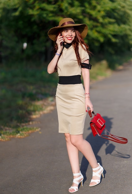 mulher feliz com um chapéu andando no parque no verão com uma bolsa e falando ao telefone